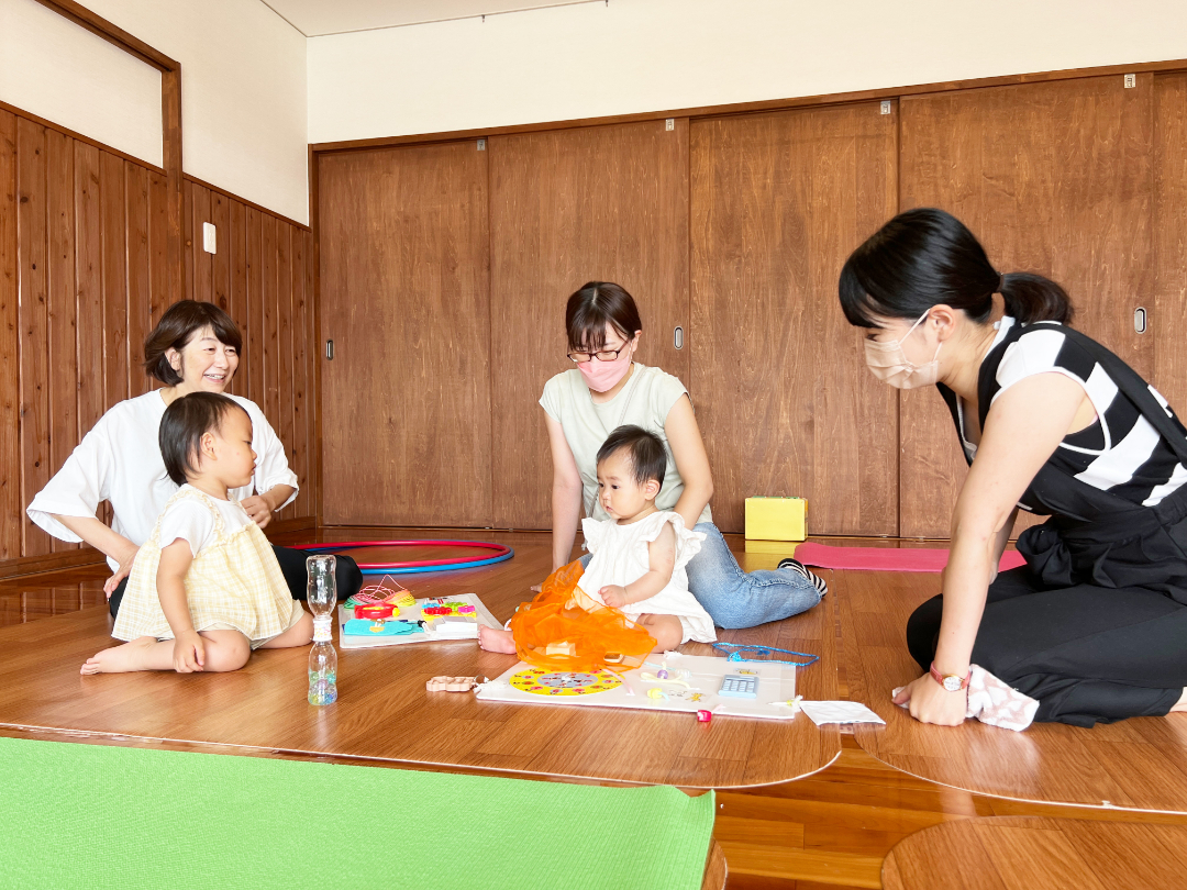 床に座り遊ぶ子供と、その周りで会話をする保育士とお母さんの写真