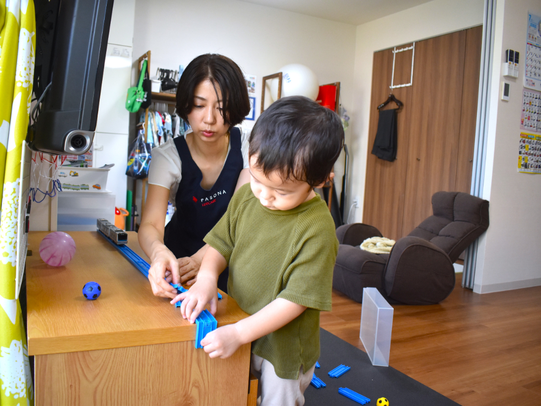 レールをつないで電車のおもちゃで遊ぶ子供とヘルパーの写真