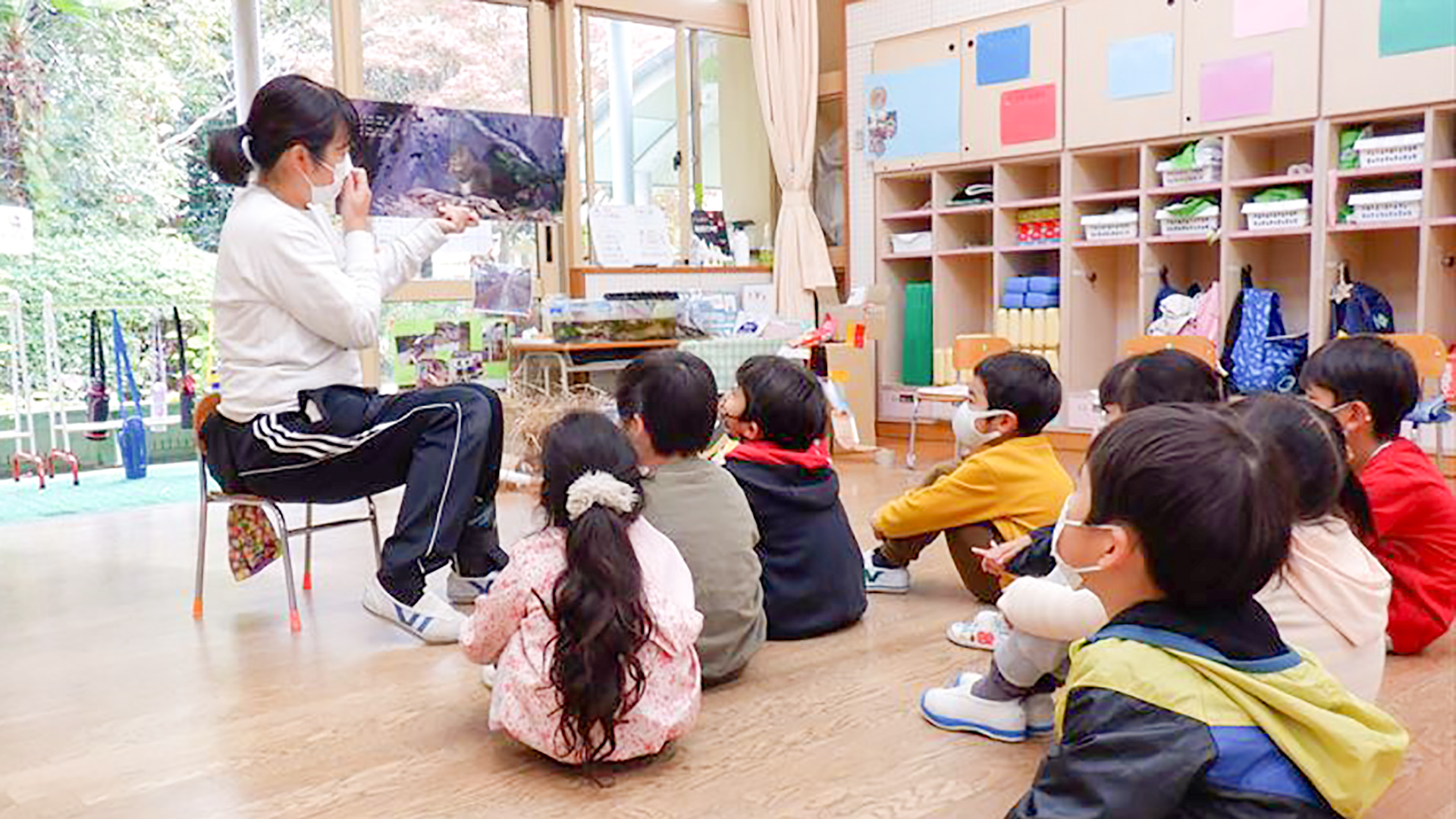 教室で先生がリスの絵本を読み聞かせしてくれています。先生の前に体育座りをして真剣に聞き入る子供たちの写真。